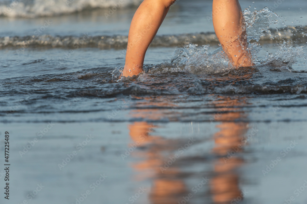 Beautiful legs of a young woman walking on a beach with water splashing against her feet