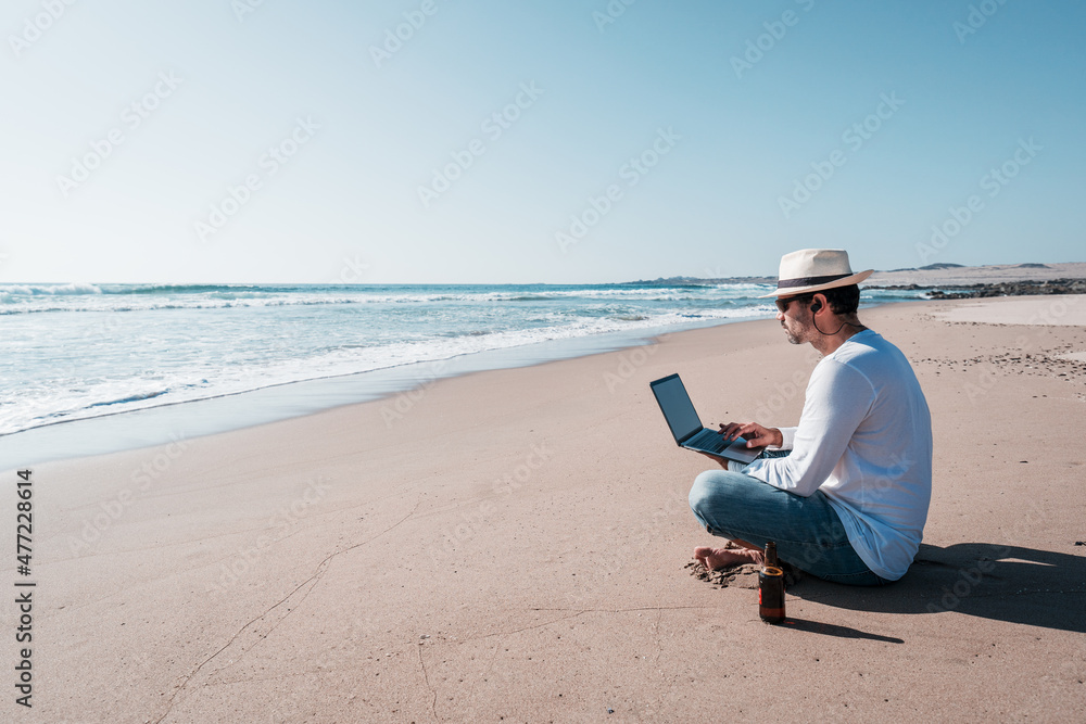 man sitting on the beach with a laptop alone doing telecommuting or remote work	and drinking beer