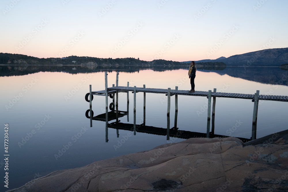 Silhouette of a young woman standing on the wooden dock at nightfall. Magical view of the lake, mountains and forest and its reflection in the lake's water. Beautiful sunset colors in the environment.