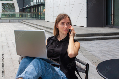 Young business woman working at the computer outside, makes the phone call on the background of modern city buildings
