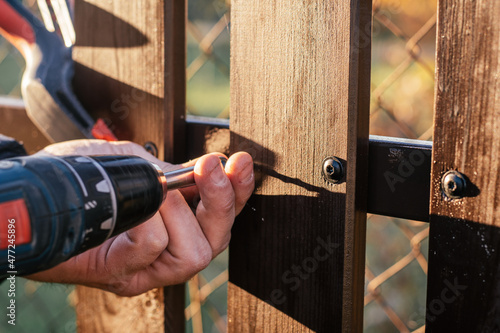 Hand of carpenter holding stud driving machine putting screw into wooden plank to metal construction for reconstructing wooden fence. Outdoor works. Chain-link fence background. Close up.