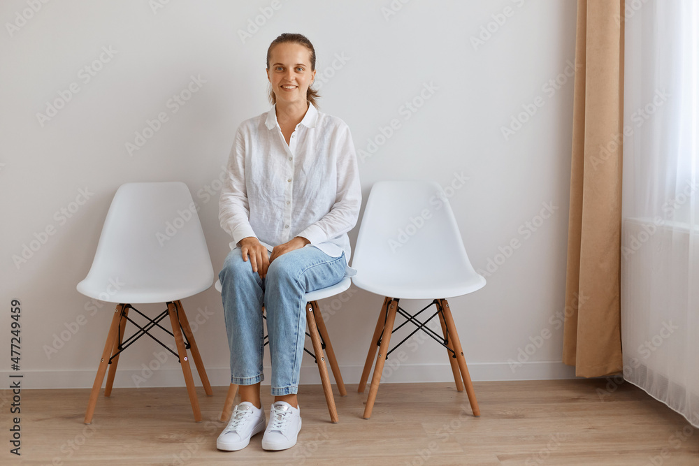 Fototapeta premium Indoor shot of attractive young adult woman sitting on chair against white wall, wearing shirt and jeans, posing with smile, looking happily at camera, expressing optimism.
