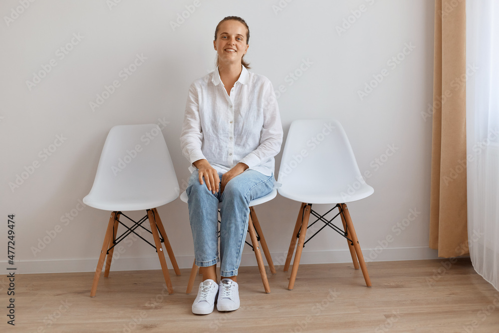 Fototapeta premium Image of smiling satisfied attractive young adult woman sitting on chair against white wall, wearing shirt and jeans, looking at camera with calm and optimistic facial expression.