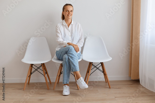 Image confident Caucasian young adult woman with ponytail wearing white shirt and jeans, sitting on chair against light wall indoor, posing with kind look and charming smile, expressing confidence.