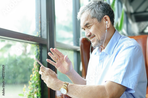 Asian senior man sitting among nature atmosphere near glasses window, talking and waving to someone by online video call on mobile smartphone(soft and selective focus)
