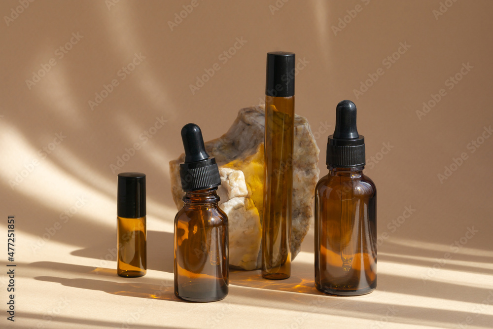 Mock-ups of glass bottles with dropper lids of various heights and shapes, standing next to marble stone. Rays of sunlight reflecting off objects create shadows on beige background