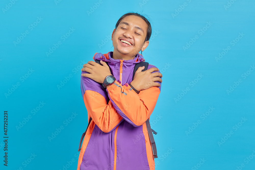 Portrait of cheerful handsome Asian man wearing jacket and carrying backpack hugging himself with both hands isolated on blue background Portrait of cheerful handsome Asian man wearing jacket and carrying backpack hugging himself with both hands isolated on blue background