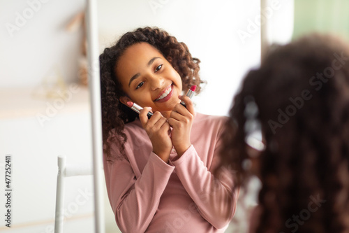 Cute african american teenage girl choosing lipstick color, looking at herself in the mirror, over the shoulder view
