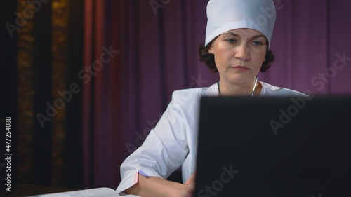 A beautiful woman doctor is working in her office at a computer. She enters the data into a laptop and then manually records the finished readings in her diary. Doctor's daily office work.
