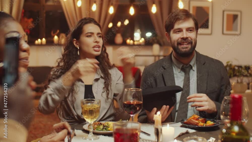 Group of modern young friends enjoying birthday party sitting at dining table playing hat word guessing game Group of modern young friends enjoying birthday party sitting at dining table playing hat word guessing game