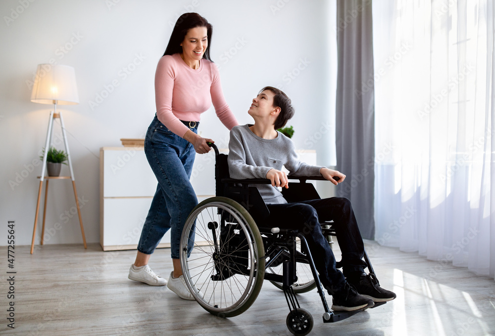 Full length portrait of young mother and her impaired son in wheelchair spending time together at home Full length portrait of young mother and her impaired son in wheelchair spending time together at home
