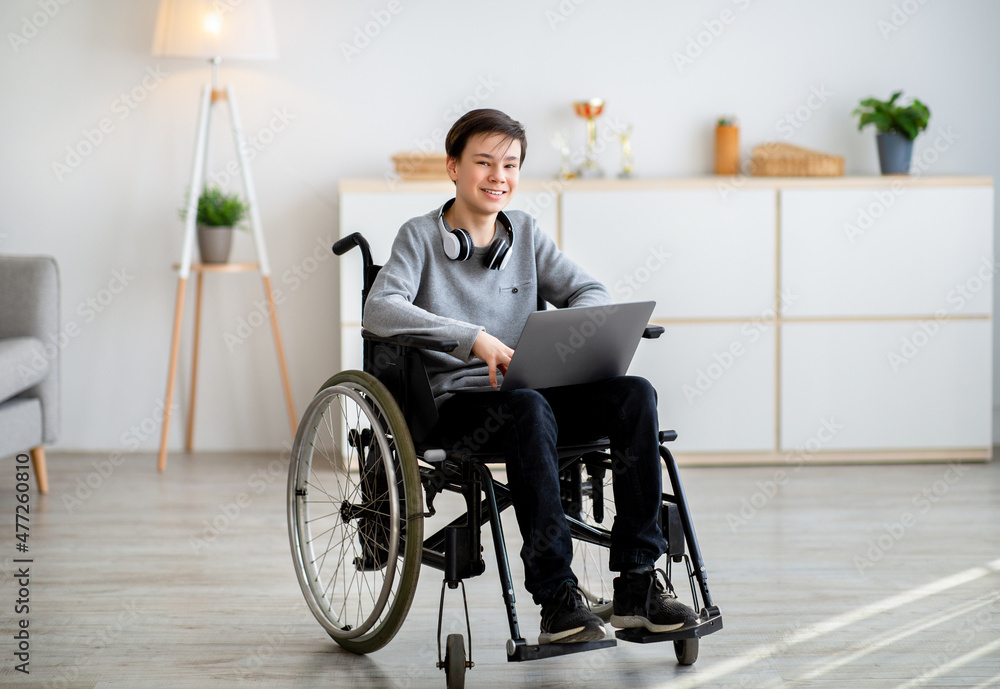 Web-based education for disabled pupils. Cheerful teen boy in wheelchair using laptop for online studies at home Web-based education for disabled pupils. Cheerful teen boy in wheelchair using laptop for online studies at home