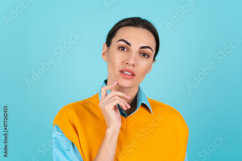 Young woman in a shirt and an orange vest on a turquoise background looks thoughtfully at the camera