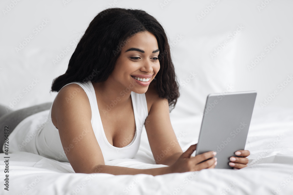 Free time and weekend pastime concept. Happy black lady holding digital tablet computer, lying on bed, free space Free time and weekend pastime concept. Happy black lady holding digital tablet computer, lying on bed, free space