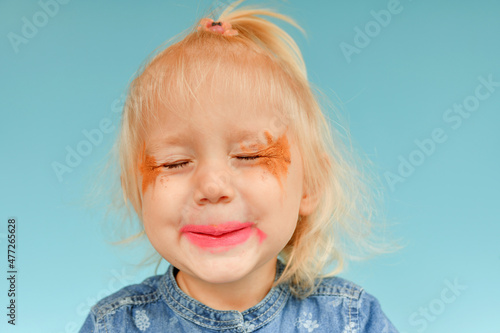 A funny and ineptly painted little girl put on her makeup and shows herself with her eyes closed.