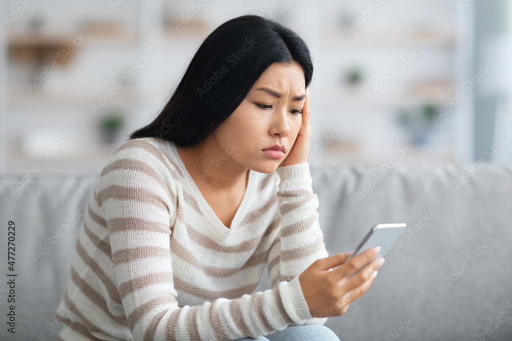 Worried Asian Lady Looking At Smartphone While Sitting On Sofa At Home Worried Asian Lady Looking At Smartphone While Sitting On Sofa At Home