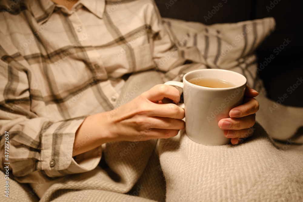 A cup of tea in the hands of a girl. The girl is drinking hot tea. A girl in pajamas is wrapped in a blanket and is drinking delicious tea. Enjoy the comforts of home. White cup close up in woman hand