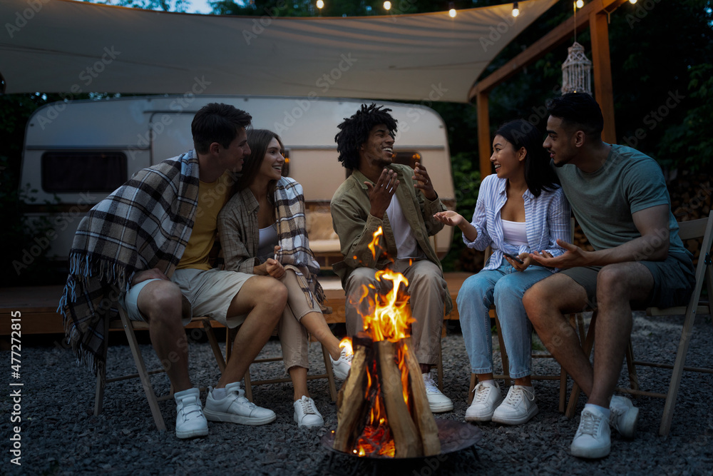 Multiethnic friends sitting beside bonfire, having fun conversation, spending time on camping trip with RV together - obrazy, fototapety, plakaty 