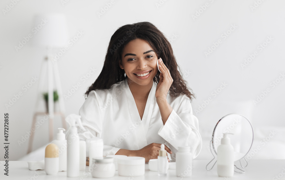 Happy african american woman removing makeup with cotton pad, sitting at dressing table in bedroom interior Happy african american woman removing makeup with cotton pad, sitting at dressing table in bedroom interior
