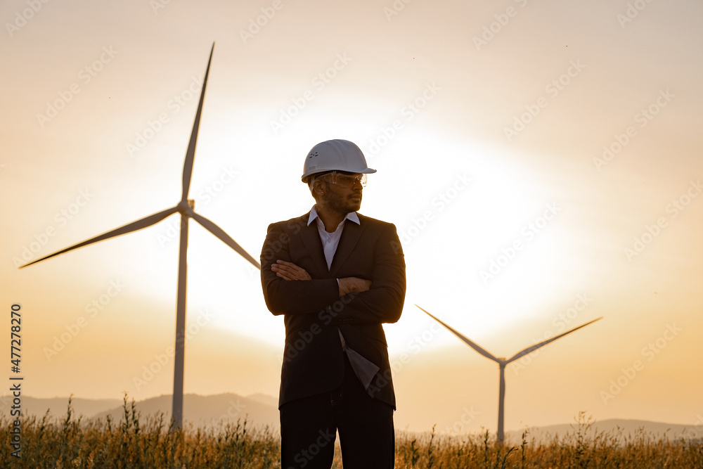 Confident indian man in suit, helmet and glasses keeping arms crossed and looking aside while standing on rural area. Background of summer sunset and huge windmills. Confident indian man in suit, helmet and glasses keeping arms crossed and looking aside while standing on rural area. Background of summer sunset and huge windmills.