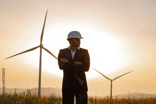 Confident indian man in suit, helmet and glasses keeping arms crossed and looking aside while standing on rural area. Background of summer sunset and huge windmills.