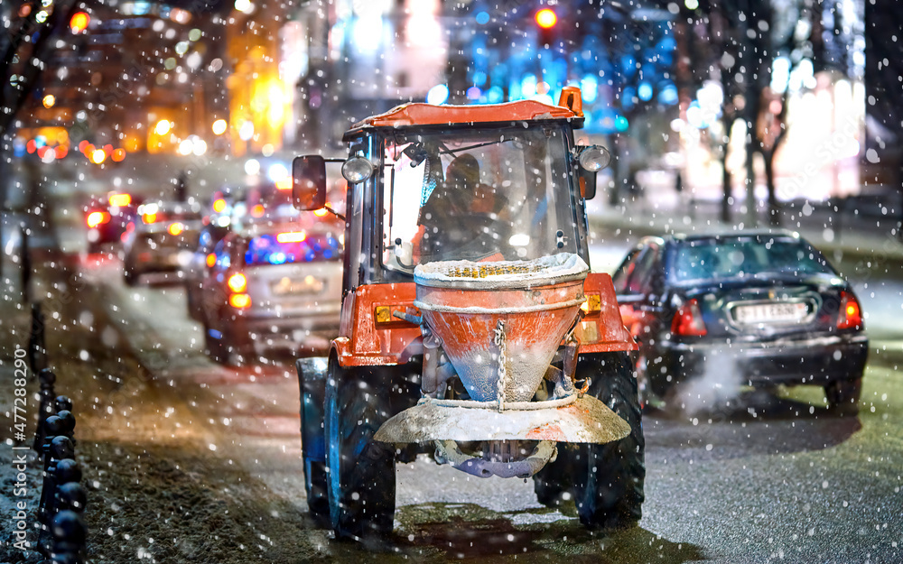 Tractor with mounted salt spreader driving on city road at night, snow removal service operating in winter during blizzard. Snow and ice removal service spread rock salt on road. Tractor with mounted salt spreader driving on city road at night, snow removal service operating in winter during blizzard. Snow and ice removal service spread rock salt on road.