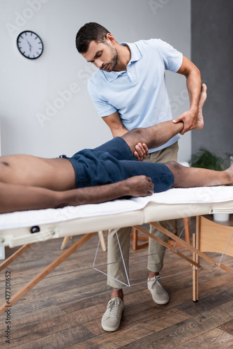 chiropractor massaging leg of african american man in rehabilitation clinic