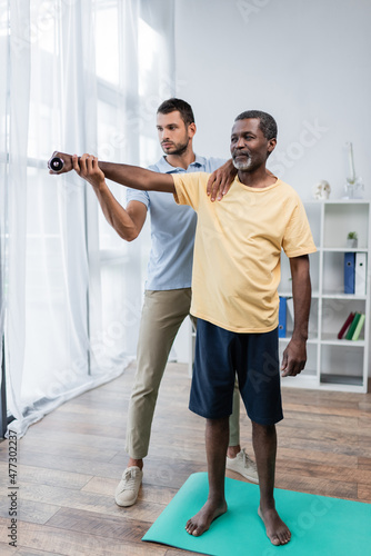 full length view of barefoot african american man exercising with dumbbell near rehabilitologist photo