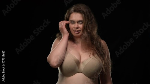 Portrait of confident overweight Caucasian woman in bra looking at camera with seductive facial expression touching hair. Self assured adult brunette lady posing at black background smiling