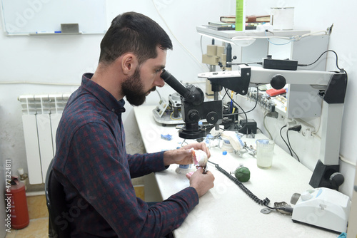 A young man looks through a microscope examining repairs dentures or a jaw in the workshop of a dental technician