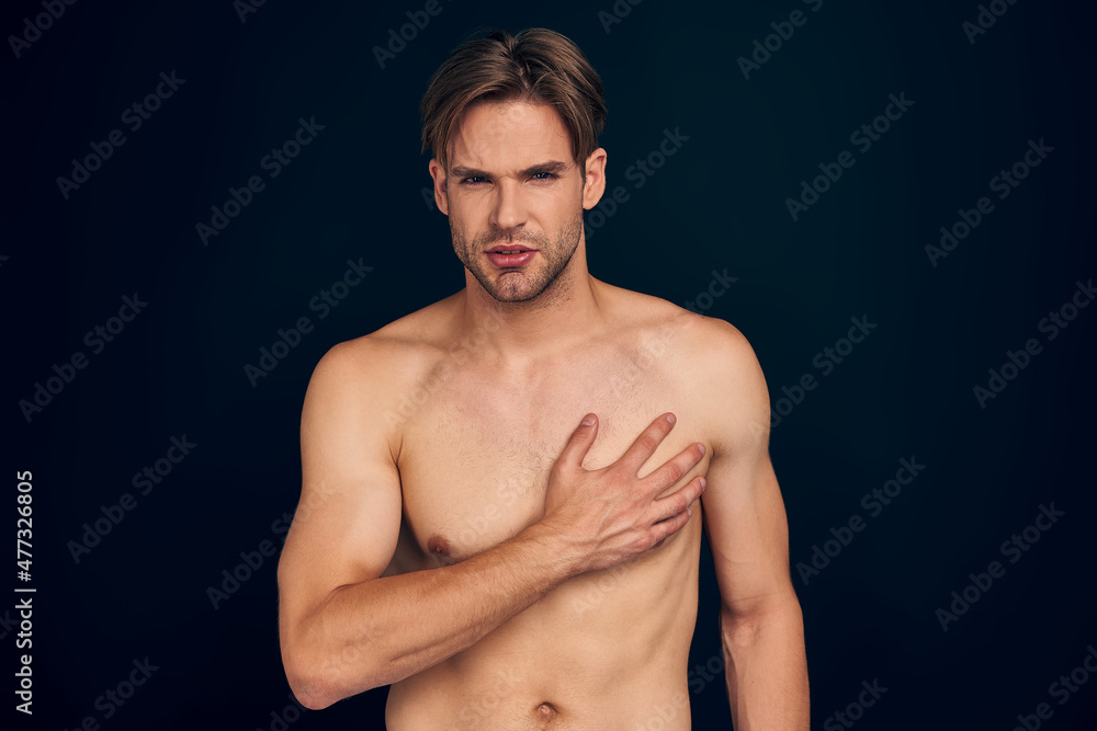 Fototapeta premium Handsome young bearded man isolated. Portrait of naked muscular man is standing on dark blue background. Man holding his heart. Experiencing heart pain