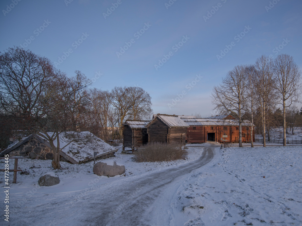 Old log stable and houses with birch-bark roofs in a snowy park a sunny winter day afternoon in Stockholm