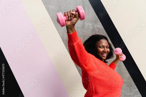 Happy plus size woman exercising with dumbbells near patterned wall