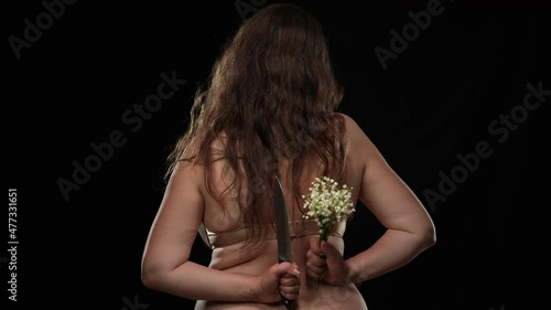 Back view plus size woman in bra holding knife and bouquet standing at black background. Obese Caucasian lady stretching hand with flowers sqeezing weapon behind. Hypocrisy and pretense concept photo