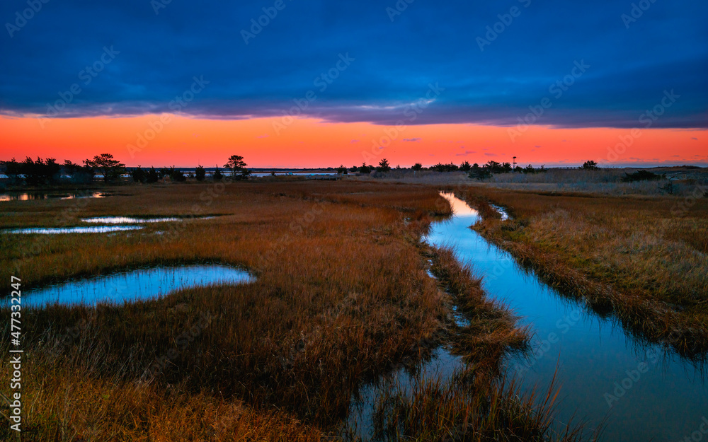 Vibrant saturated sunrise seascape over the marsh river in Mashpee, Massachusetts. Blue clouds, pink sky, brown grasses, and turquoise-colored seawater reflections.