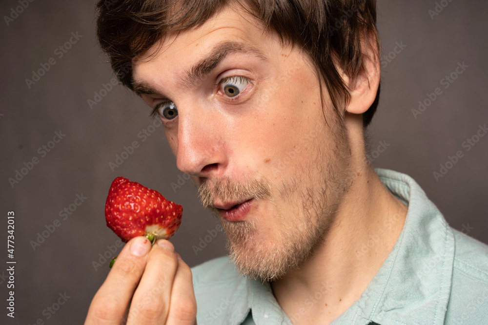 Young handsome tall slim white man with brown hair holding half eaten strawberry making a silly face in light blue shirt on grey background Young handsome tall slim white man with brown hair holding half eaten strawberry making a silly face in light blue shirt on grey background