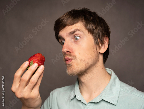 Young handsome tall slim white man with brown hair hypnotizing strawberry in light blue shirt on grey background