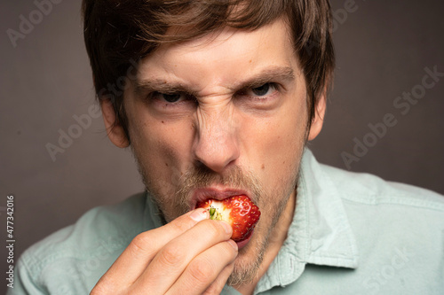 Young handsome tall slim white man with brown hair angrily biting strawberry in light blue shirt on grey background