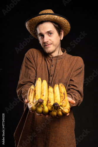 Young handsome tall slim white man with brown hair offering bunch of bananas with brown shirt and straw hat on black background