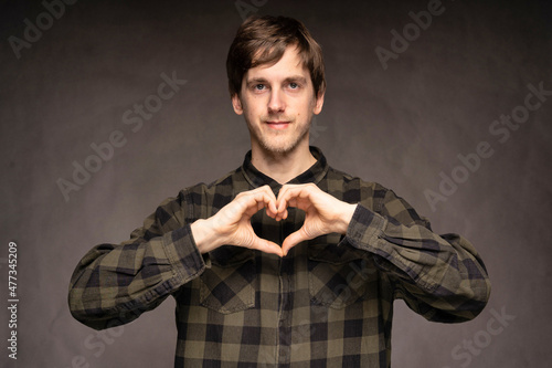 Young handsome tall slim white man with brown hair making heart with hands with flannel shirt on grey background
