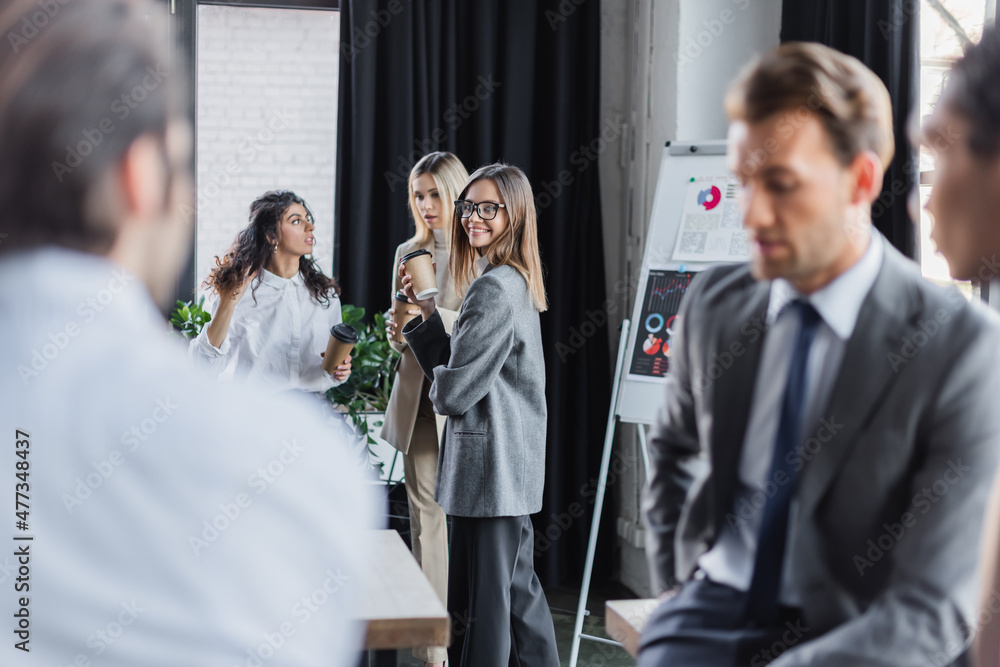 Fototapeta premium happy woman holding coffee to go near colleagues and blurred multiethnic businessmen.