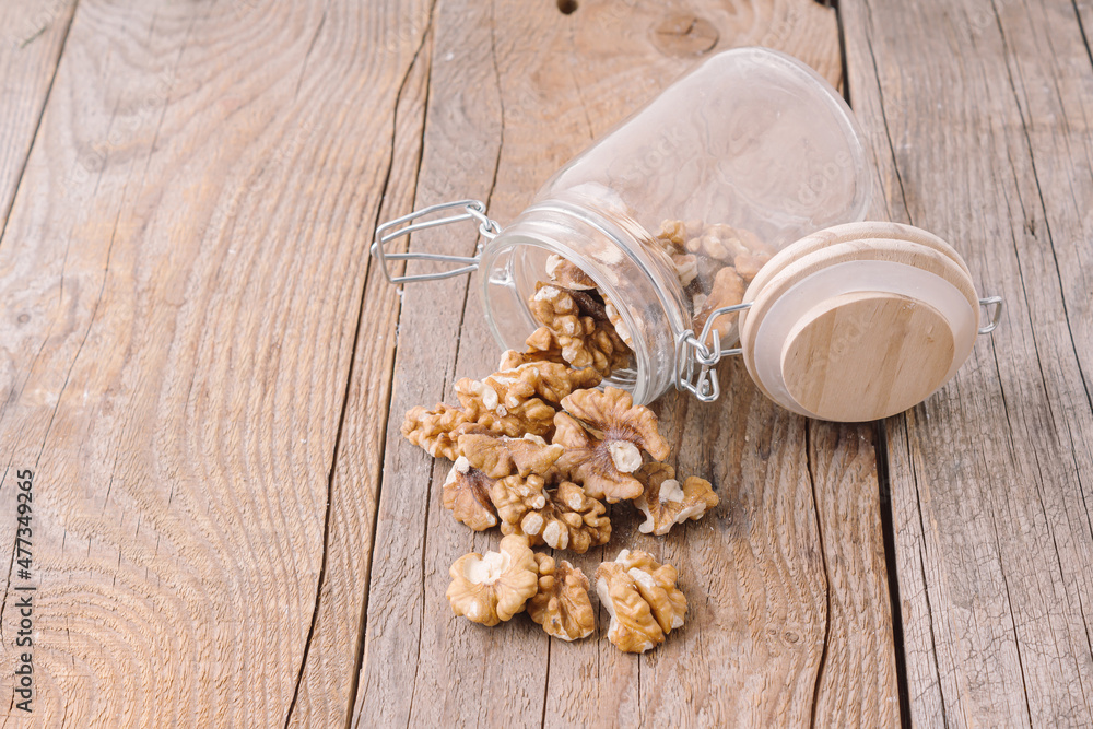 Fototapeta premium Peeled walnuts in glass jar on wooden background. Storage of nuts. Storage container at kitchen. Scattered walnuts. Space for text