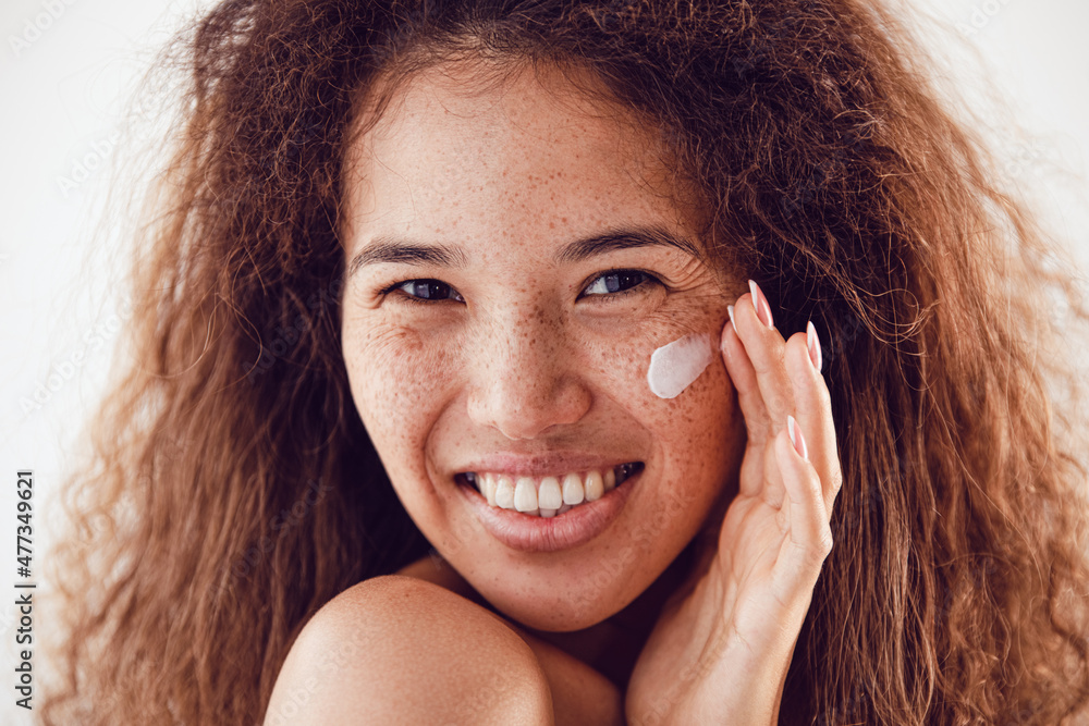 Portrait of woman with curly hair and freckles applying moisturizer to her face. Portrait of woman with curly hair and freckles applying moisturizer to her face.