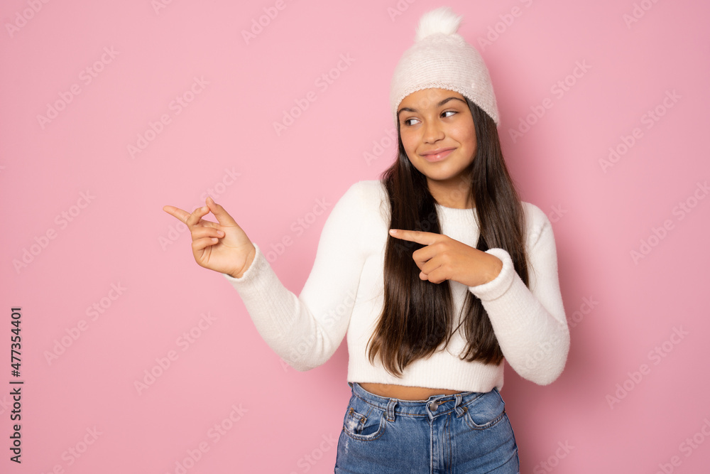 Young smiling girl wearing winter hat over isolated pink background pointing fingers to the side Young smiling girl wearing winter hat over isolated pink background pointing fingers to the side