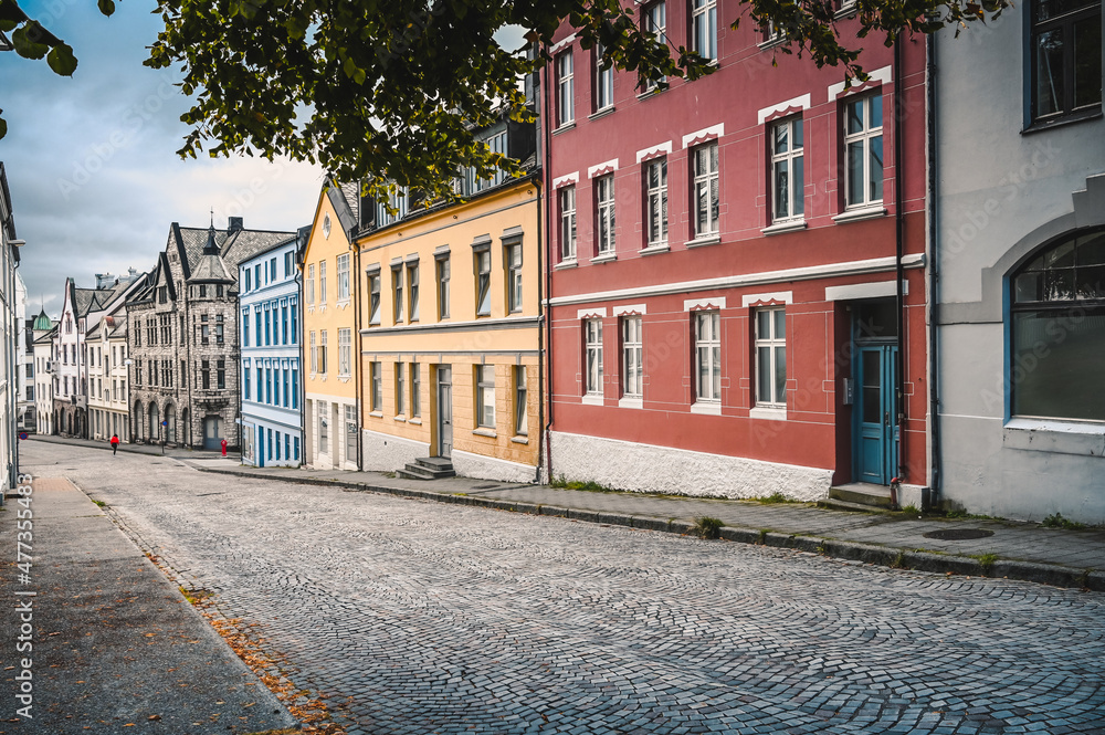 Empty street with colorful houses in the city of Alesund, Norway. Leaves of a tree are framing the picture. No people are on the road due to the Corona pandemic. 