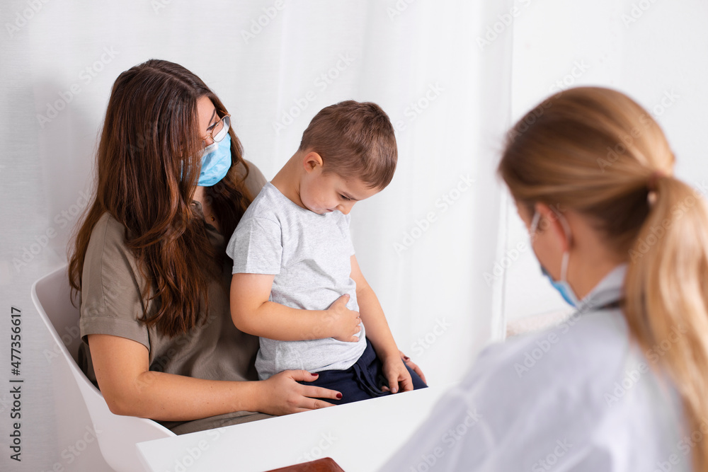 Obraz premium Young brunette woman with little boy having consultation at pediatrician office. Child has abdominal pain. Doctor, child and mother wearing facemasks during coronavirus and flu outbreak. Covid 19.