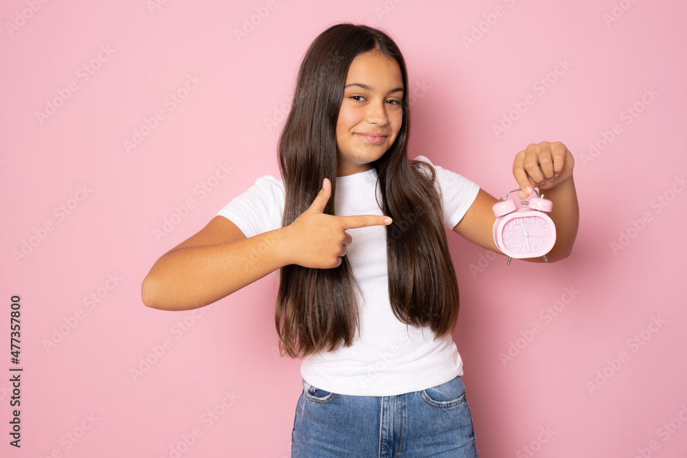 Beautiful smiling girl wearing casual white t-shirt showing alarm clock standing isolated over pink background. Beautiful smiling girl wearing casual white t-shirt showing alarm clock standing isolated over pink background.