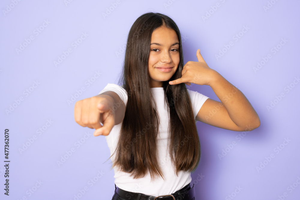 Beautiful girl wearing white t-shirt standing over purple isolated background smiling doing talking on the telephone gesture and pointing to you. Call me. Beautiful girl wearing white t-shirt standing over purple isolated background smiling doing talking on the telephone gesture and pointing to you. Call me.