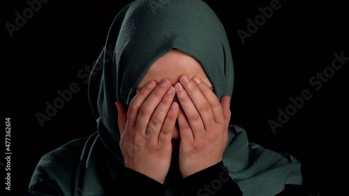 Close up of a sad muslim girl's face covering her face with her hands. Isolated on a black background.