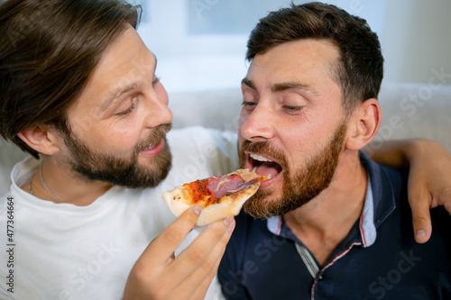 Gay marriage. The guys are in the living room, sitting on the couch with pizza. They give each other a slice of pizza. Both young and European.